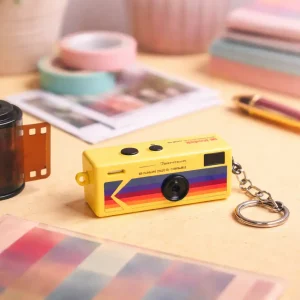 Mini cámara digital tipo llavero de estilo retro colocada sobre una mesa con elementos fotográficos vintage y luz cálida
