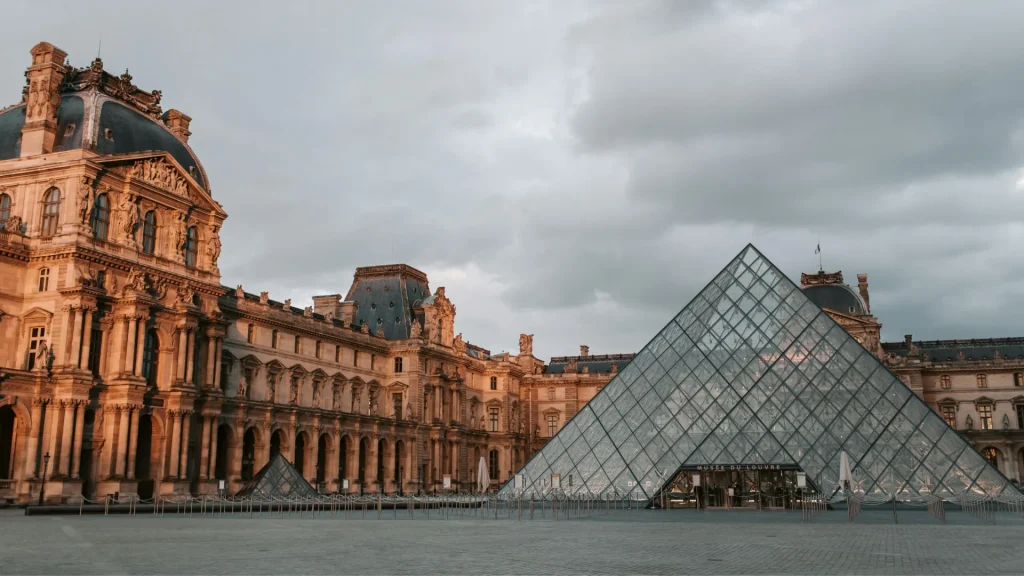 Pirámide de cristal del Louvre Museum en París frente al histórico palacio bajo un cielo nublado.