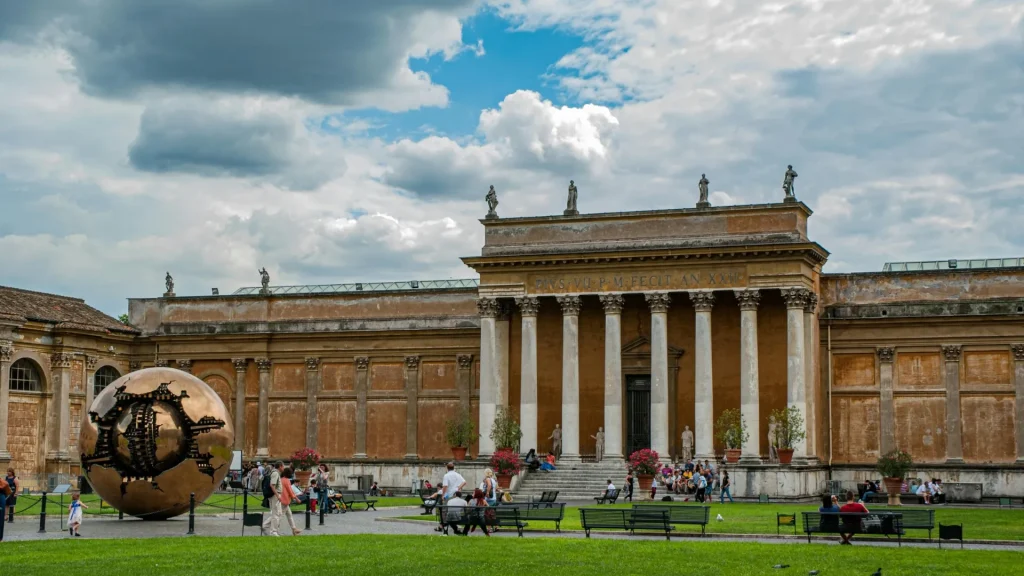 Patio de los Vatican Museums con edificio clásico, columnas y visitantes bajo cielo nublado.