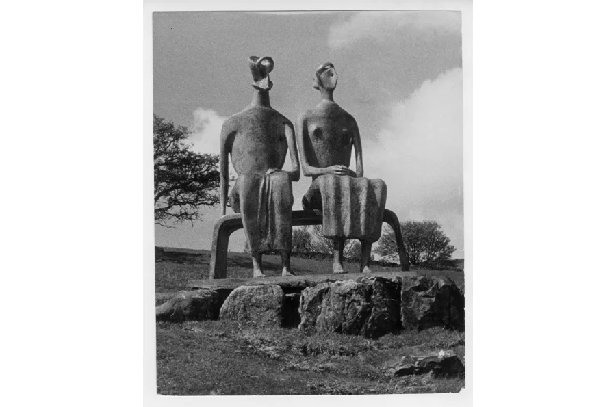 Fotografía histórica en blanco y negro de la escultura King and Queen de Henry Moore, sentada sobre un banco en un paisaje natural.